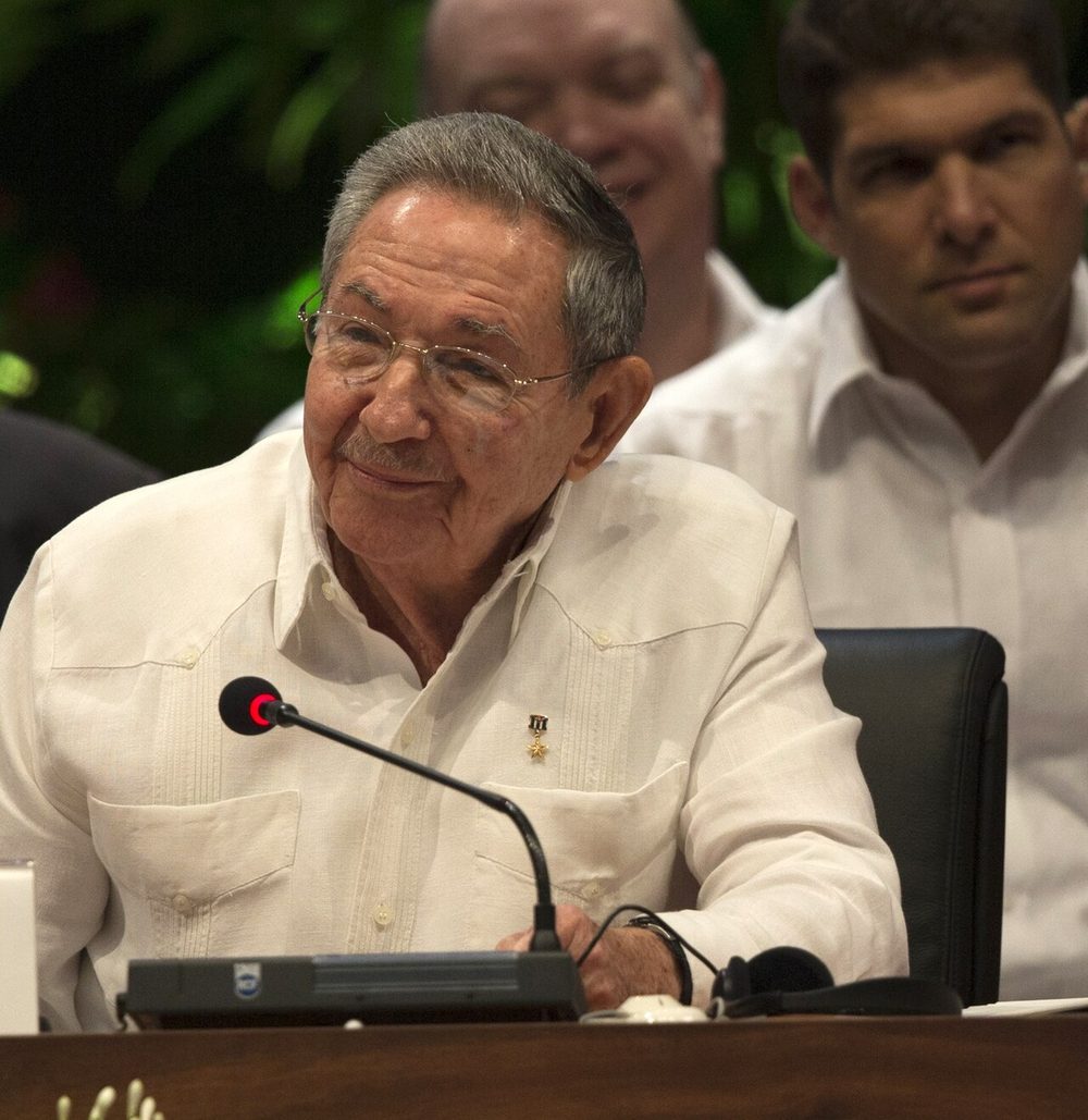 Raul Castro in camicia bianca, seduto a un tavolo di conferenza davanti a un microfono, sorride guardando di lato