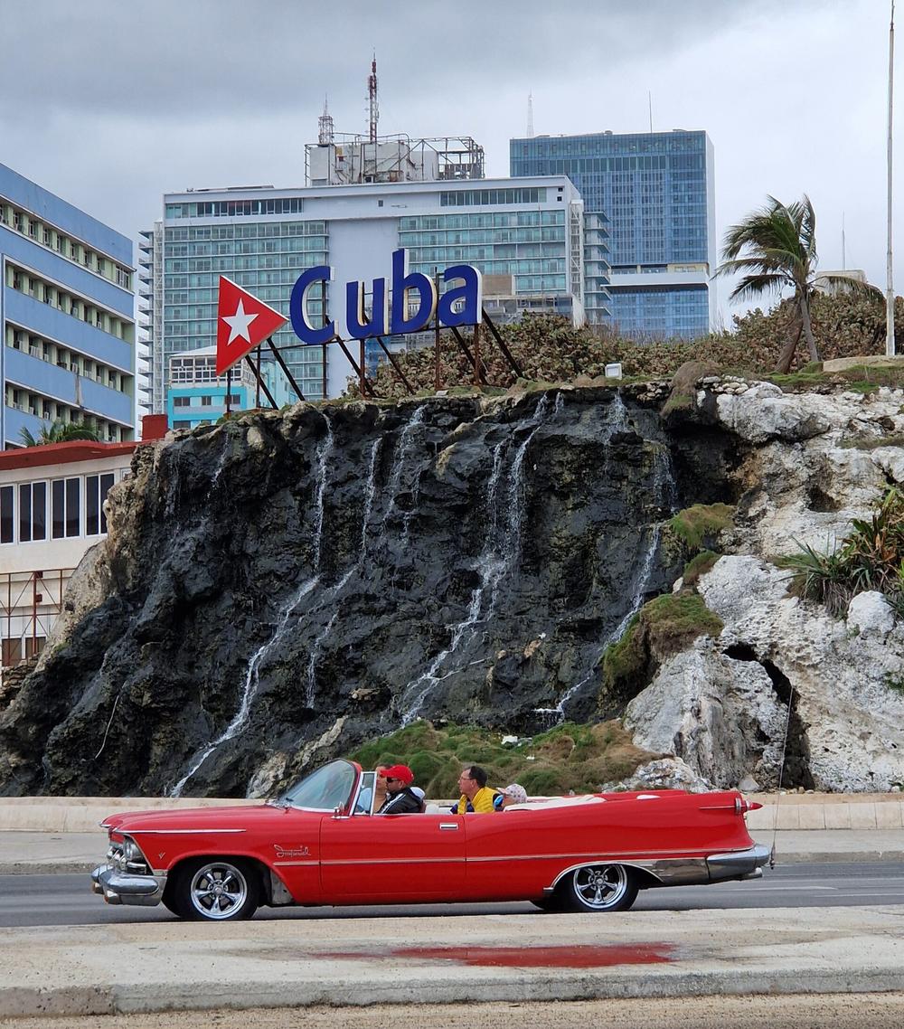 Il Malecon dell'Avana: in primo piano un'auto d'epoca rossa e l'insegna Cuba su una roccia; dietro, l'hotel Habana Libre e, a destra, la torre di vetro della Torre K